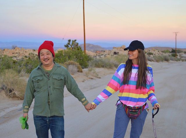 Khalyla Kuhn and Bobby Lee holding each other's hands. Bobby Lee wearing a red cap, mint green shirt and a blue jeans. Khalyla Kuhn in a black cap, colorful t-shirt and blue jeans.